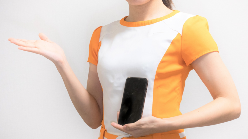 イベントコンパニオンがスマートフォンを左手に持ち、右手で紹介をする手振りをしている様子